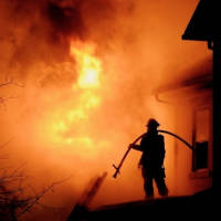 Firefighter Silhouetted Against Fiery Background Firefighter holding a hose over his shoulder silhouetted against a fiery background.